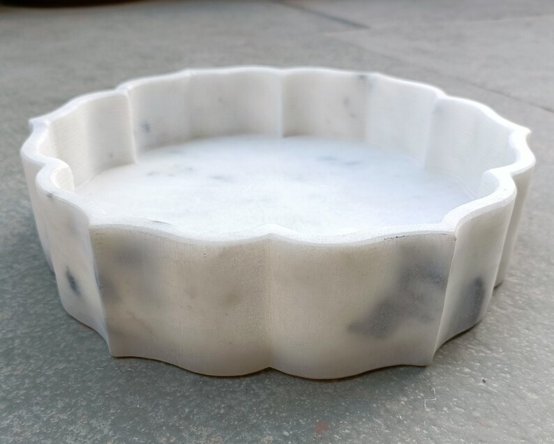 Puede incluir: Bandeja decorativa redonda de m&aacute;rmol blanco con borde festoneado. La bandeja es lisa y pulida.