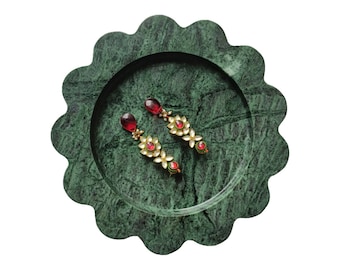May include: A pair of gold-toned earrings with red gemstones and white floral accents rests on a green marble tray. The earrings have a teardrop-shaped red stone at the top and a series of smaller red stones and white floral designs.