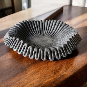 Könnte beinhalten: Eine graue Steinschüssel mit einem gewellten Rand. Die Schüssel ist blumenförmig und hat einen glatten, polierten Innenraum.