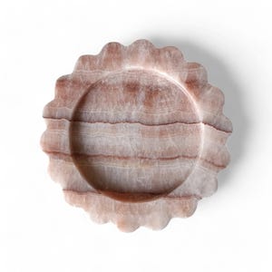 May include: A circular tray carved from pink and white banded stone, featuring a scalloped edge and a smooth, recessed center. The natural mineral patterns create horizontal layers of varying shades of light and dark dusty rose.