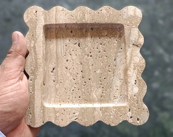 May include: A decorative tray made of light brown travertine stone with a wavy edge. The tray has a rectangular inset and a textured surface.