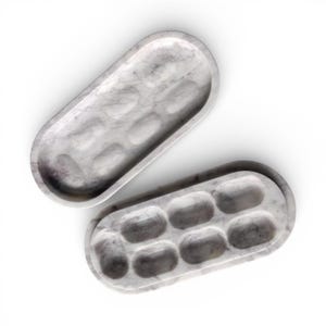 Puede incluir: Dos bandejas ovaladas de mármol con un patrón veteado gris y blanco. Cada bandeja tiene seis compartimentos empotrados. Las bandejas están diseñadas para organizar objetos pequeños y tienen un acabado liso y pulido.