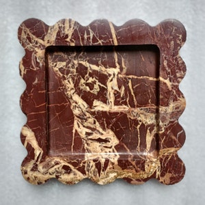 May include: A decorative tray made of red and cream marble with a scalloped edge. The tray has a rectangular shape with a raised rim.