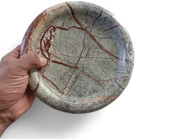 May include: A circular stone bowl featuring a polished surface with a mottled sage green base color and prominent, intersecting dark red veins running throughout the material, creating a natural, marbled appearance across the entire object.