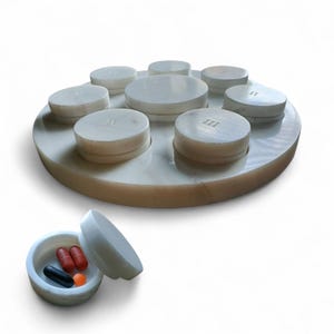 Pode incluir: Um organizador de comprimidos circular em mármore branco com sete compartimentos individuais com tampa dispostos numa base. Cada tampa está gravada com numerais romanos de I a VII. Um compartimento aberto separado mostra dois comprimidos vermelhos, um preto e um laranja.