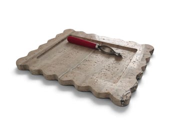 May include: A rectangular beige travertine stone serving board with a scalloped edge design. Resting on the textured surface is a metal tool featuring a bright red handle, designed with a pointed tip and a circular opening for food preparation.