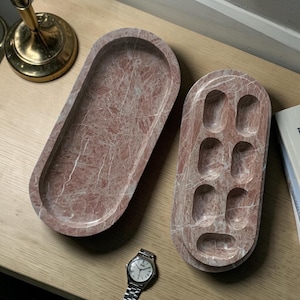 May include: Two oval-shaped, pink marble trays with white veining. One tray is a simple, open design, while the other has six recessed compartments. A silver wristwatch with a white face is next to the trays.