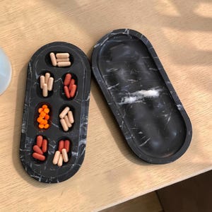 May include: Two oval-shaped black marble trays with white veining. One tray features six recessed compartments containing various beige, orange, and reddish-brown capsules and round tablets. The second tray is empty with a smooth, solid surface.