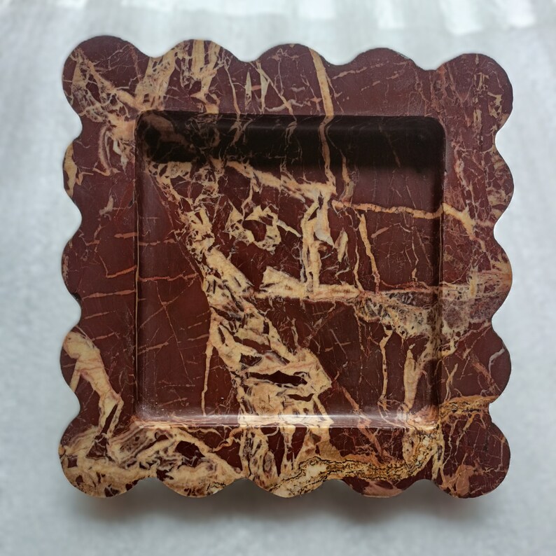 May include: A decorative tray made of red and cream marbled stone with a scalloped edge. The tray has a rectangular shape and a raised rim.
