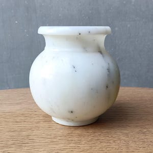 May include: A white marble vase with black speckles. The vase has a round body and a slightly flared opening.