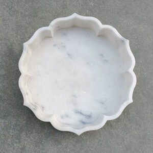 Puede incluir: Un cuenco decorativo de m&aacute;rmol blanco con un borde festoneado. El cuenco es redondo y tiene una superficie lisa y pulida.