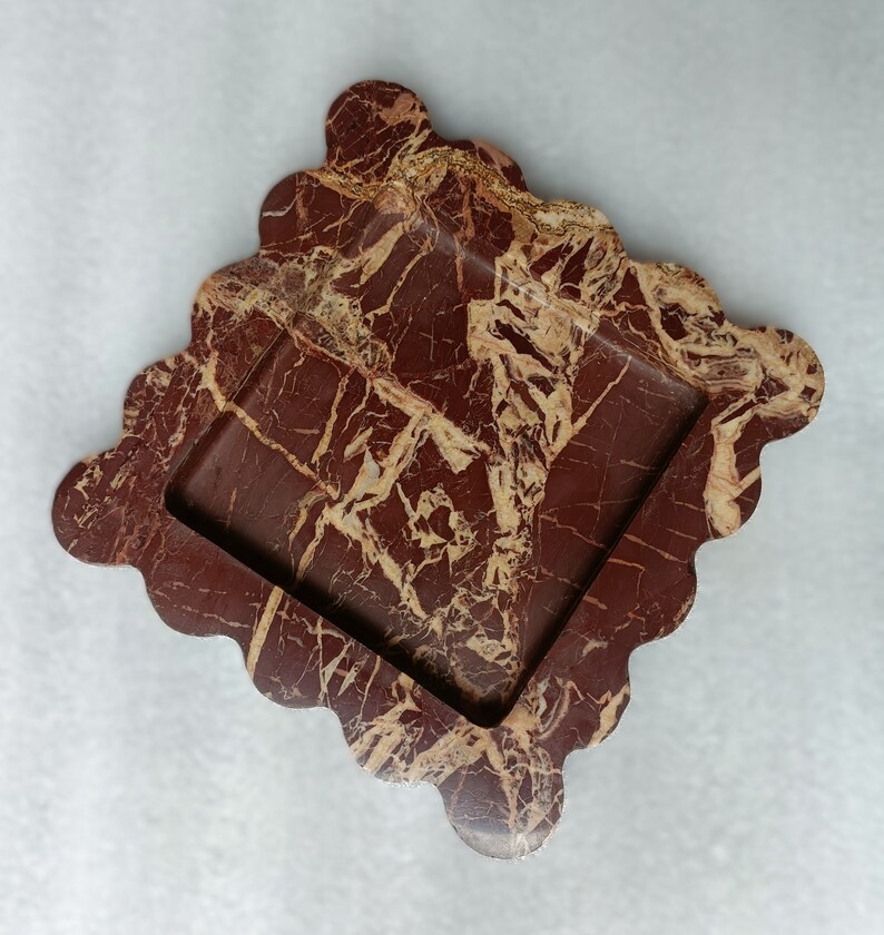 May include: A square-shaped, red and gold marble tray with a wavy edge. The tray has a square inset in the center.