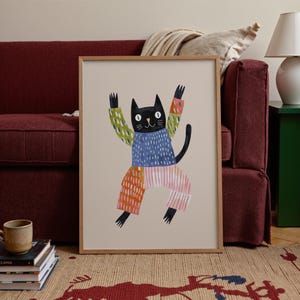 Puede incluir: Una impresión artística enmarcada presenta una ilustración caprichosa de un gato negro con los brazos levantados. El gato lleva un suéter azul, pantalones coloridos y una manga estampada. La obra de arte está enmarcada en madera clara sobre un fondo neutro.