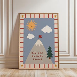 Peut inclure: Une affiche encadrée avec un message de motivation. L'œuvre représente une montagne avec un drapeau rouge, un soleil souriant, des nuages et un arbre vert sur fond bleu. Le texte dit "You can do hard things."