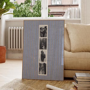 May include: A rectangular photo frame with a blue and white striped fabric background. The frame holds four black and white photographs. The frame is leaning against a beige sofa, with a window and plant in the background.