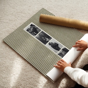 May include: A striped, olive green and white photo mat with three black and white photographs. A rolled-up brown paper tube rests on the mat. A person is unrolling a white paper.