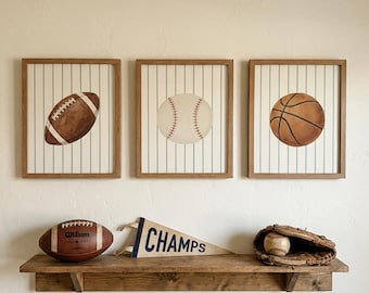May include: Three framed sports-themed prints: a football, a baseball, and a basketball, each set against a striped background. Below, a wooden shelf displays a football, a pennant reading "CHAMPS," and a baseball glove with a ball.