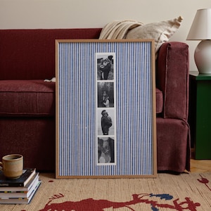May include: A framed photo display with a blue and white striped fabric background. The frame holds four black and white photographs. The frame is made of light-colored wood. The display is in front of a burgundy sofa.
