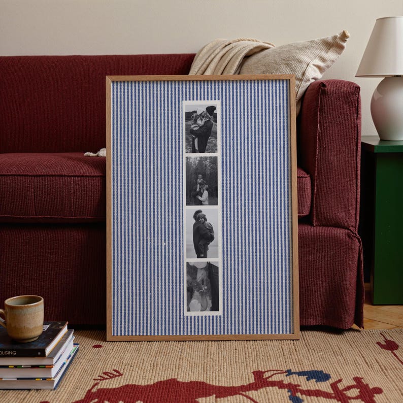 May include: A framed photo display with a blue and white striped fabric background. The frame holds four black and white photographs. The frame is made of light-colored wood. The display is in front of a burgundy sofa.