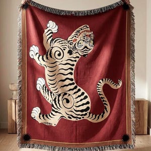 Puede incluir: Una manta tejida roja con un diseño de tigre crema y negro. La manta tiene un borde con flecos y un borde marrón. El tigre se representa en una pose dinámica, con la boca abierta y las patas levantadas. Ideal para un sofá.