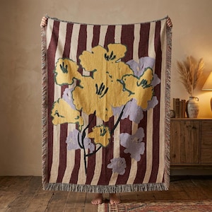 May include: A woven throw blanket with a floral design. The blanket has vertical burgundy and cream stripes, with large yellow and lavender flowers. The edges have a fringed trim.