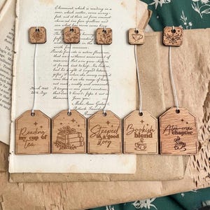 May include: Five wooden tea bag tags with white strings and small square wooden toppers. Each tag features laser-engraved illustrations and text including Reading my cup of tea, Steeped in a good story, Bookish blend, and A tea-rific chapter.
