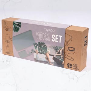 Puede incluir: Caja rectangular de cartón con la inscripción "my yoga YOGA SET". Contiene una esterilla de yoga, un bloque y una correa. También hay ilustraciones de posturas de yoga y el texto "PLANET FRIENDLY PRODUCTS".
