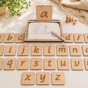 May include: A wooden alphabet learning set with a tray for tracing letters in sand. Includes individual letter tiles and a wooden stylus. The letter "a" is traced in the sand. Designed for early childhood education and learning.