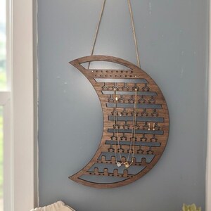 May include: A wooden crescent moon-shaped jewelry organizer with multiple slots and holes for earrings and necklaces. The organizer is hanging on a light blue wall. A small bag and a potted plant are on a wooden surface below.