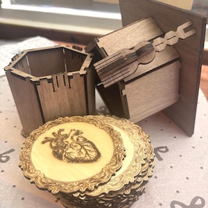 Peut inclure: Ensemble d'objets en bois découpés au laser. Comprend un récipient hexagonal, une petite boîte avec une poignée décorative et une pile de dessous de verre ronds. Les dessous de verre présentent un motif de cœur anatomique gravé dans une bordure ornée. Les objets sont en bois brun clair.