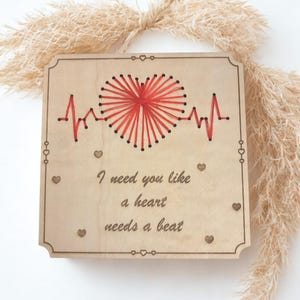 May include: A wooden plaque with a red heart-shaped string art design and a red heartbeat line. The text "I need you like a heart needs a beat" is engraved on the plaque. Decorative hearts are placed around the edges.