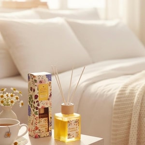 Può includere: Un diffusore in vetro giallo con bastoncini e tappo in legno è posto su un tavolo, accanto a una scatola a motivi floreali. Sul tavolo sono presenti anche una tazza di tè e un piccolo vaso di fiori. Atmosfera da camera da letto.