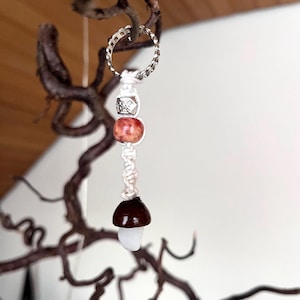 Puede incluir: Un llavero artesanal con forma de seta con un anillo plateado. El llavero presenta un cordón blanco, una cuenta marrón rojiza, una cuenta plateada y un dije de seta con un sombrero marrón y un tallo blanco. Cuelga de una rama retorcida.