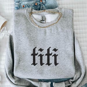 May include: A gray sweatshirt with the word "titi" in black gothic lettering, paired with blue denim jeans and white high-top sneakers. A gold chain necklace is draped on the sweatshirt. The items are arranged on a white, textured surface.