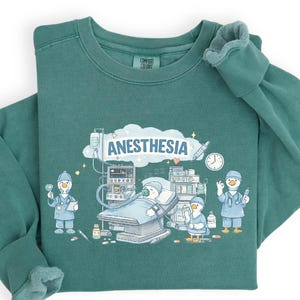 May include: Teal-colored sweatshirt with a graphic featuring cartoon ducks in medical attire, operating room equipment, and the word "ANESTHESIA" in a cloud. The design includes a clock, medical bottles, and a patient on a bed.