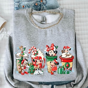 May include: A gray sweatshirt featuring a colorful graphic of Disney characters in holiday-themed coffee cups. The image includes white high-top sneakers and a pair of light-wash denim shorts. A gold chain is visible at the neckline of the sweatshirt.