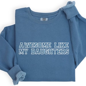 以下が含まれることがあります： 「AWESOME LIKE MY DAUGHTERS」の文字が白でプリントされた青いスウェットシャツ。長袖シャツはきれいに折りたたまれ、袖と首元が見えます。生地は柔らかく快適に見えます。