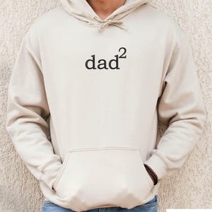May include: Sand-colored hoodie with a front pocket and a drawstring hood. The word "dad²" is printed in black on the front. The word "SAND" is printed in a white rectangle at the bottom of the image. The hoodie is made of a soft material.