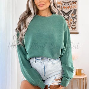 May include: A teal green crewneck sweatshirt with long sleeves. The text "Unique Blank Sweatshirt" is printed on the front. The model is wearing light blue denim shorts. The background includes a wall with a butterfly print.
