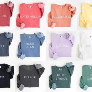 May include: A collection of long-sleeved sweatshirts in various colors, including crimson, watermelon, terracotta, and butter. Each shirt displays a color name in white text. The sweatshirts have a relaxed fit and cuffed sleeves.