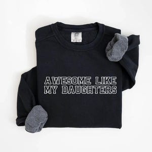 以下が含まれることがあります： 「AWESOME LIKE MY DAUGHTERS」の白い文字がプリントされた黒いスウェットシャツ。長袖シャツは灰色の袖口で、白い背景に表示されています。快適でカジュアルなスタイルです。
