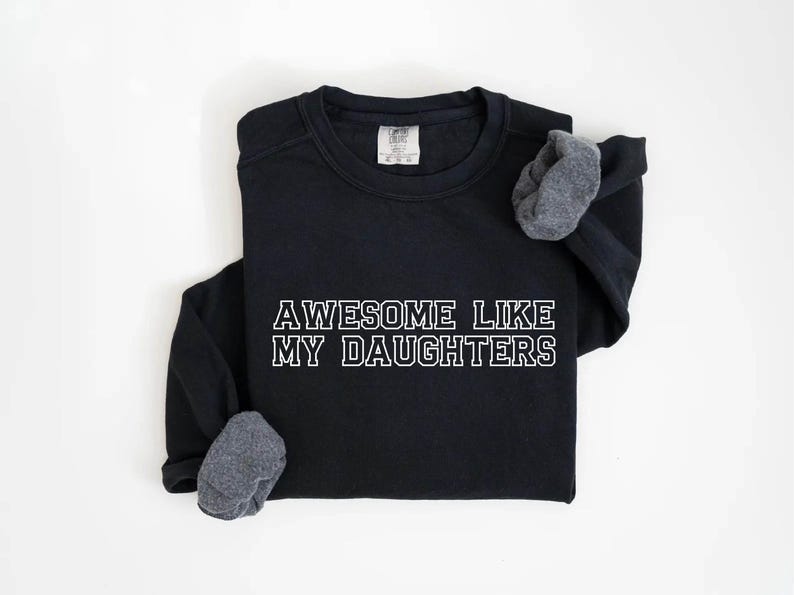 以下が含まれることがあります： 「AWESOME LIKE MY DAUGHTERS」の白い文字がプリントされた黒いスウェットシャツ。長袖シャツは灰色の袖口で、白い背景に表示されています。快適でカジュアルなスタイルです。