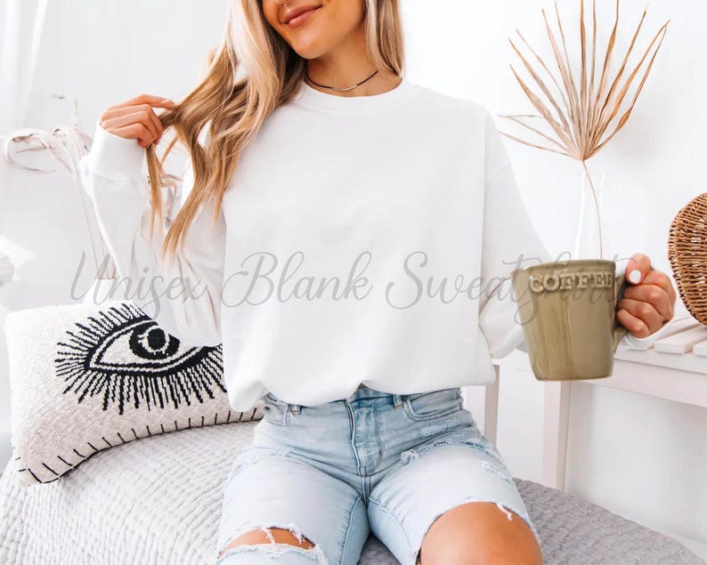 May include: A white crew neck sweatshirt with the text "Unisex Blank Sweatshirt" in a script font. The person is holding a green mug that says "Coffee". The person is wearing light blue denim shorts.