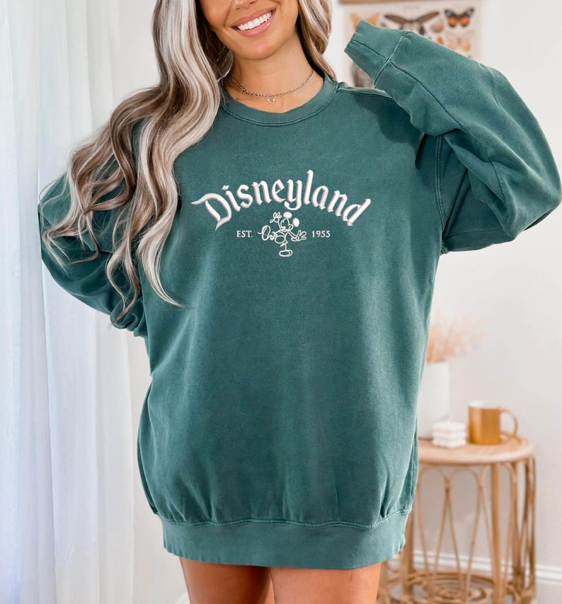 Puede incluir: Sudadera de color verde azulado con la palabra "Disneyland" en escritura blanca en el pecho. Debajo de la palabra "Disneyland" est&aacute; el texto "EST. 1955" y un peque&ntilde;o gr&aacute;fico de Mickey Mouse. La sudadera tiene mangas largas y cuello redondo.