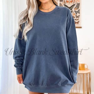 May include: A faded blue unisex sweatshirt. The garment has a crew neck and long sleeves. The text "Unisex Blank Sweatshirt" is printed on the front. The sweatshirt is a solid color and appears to be made of a soft material.