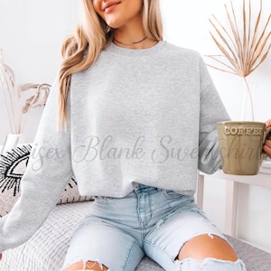 May include: A light gray crewneck sweatshirt with the text "isex Blank Sweatshirt" in a script font. The person is holding a green mug that says "COFFEE". The person is wearing light blue jeans.