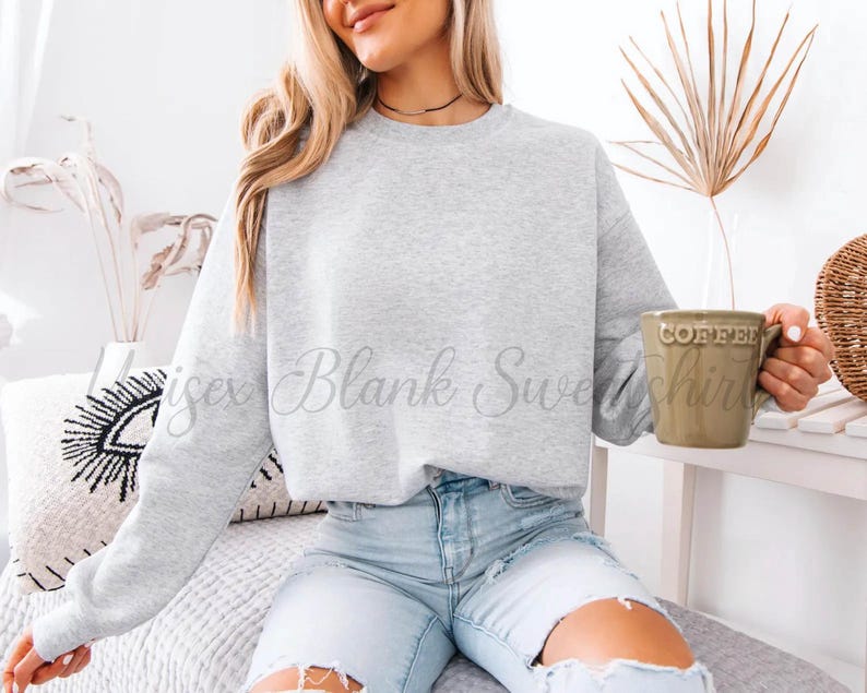 May include: A light gray crewneck sweatshirt with the text "isex Blank Sweatshirt" in a script font. The person is holding a green mug that says "COFFEE". The person is wearing light blue jeans.