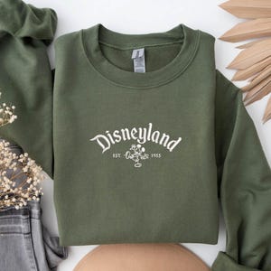 Puede incluir: Una sudadera verde oliva con la palabra "Disneyland" en escritura blanca. Debajo, un gr&aacute;fico de Mickey Mouse y el texto "EST. 1955". La sudadera est&aacute; doblada, con las mangas visibles. El fondo incluye elementos decorativos.