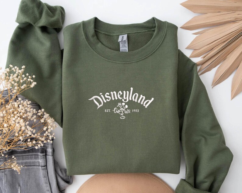 Puede incluir: Una sudadera verde oliva con la palabra "Disneyland" en escritura blanca. Debajo, un gr&aacute;fico de Mickey Mouse y el texto "EST. 1955". La sudadera est&aacute; doblada, con las mangas visibles. El fondo incluye elementos decorativos.