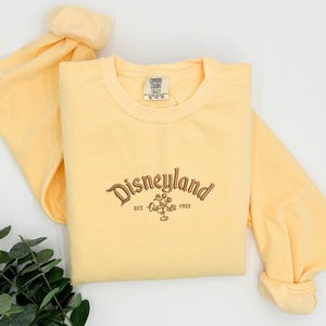 Puede incluir: Una sudadera amarilla clara con la palabra "Disneyland" bordada en escritura marr&oacute;n, junto con un gr&aacute;fico de Mickey Mouse y el a&ntilde;o "1955". La camisa de manga larga est&aacute; hecha de un material suave.
